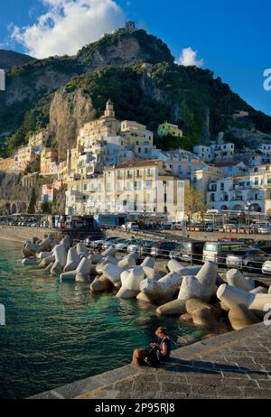 Person sitting on the main jetty at Amalfi, Salerno, Italy  with the town rising, clinging to the hillside. Amalfi Coast. Tyrrhenian Sea. Stock Photo