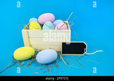 Easter moc-up. Wooden box with straw and colorful eggs on a blue background. Stock Photo