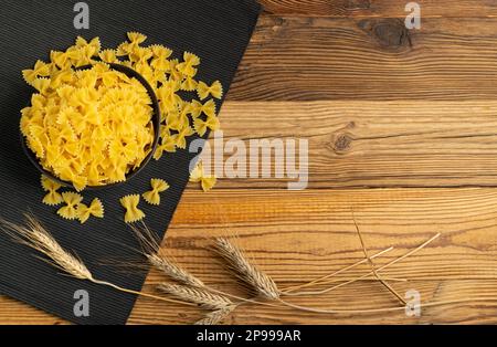 Raw Farfalle Pasta on Wooden Table, Yellow Dry Butterfly Noodles, Wheat Bow Macaroni, Uncooked Farfalle in Black Bowl on White Background Top View, Co Stock Photo