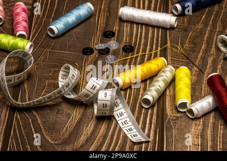 a sewing still life with a plastic ruler, needles, colored buttons, tape measure, assorted threads, golden vintage scissors on a brown wooden table Stock Photo
