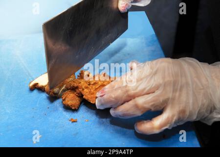 Chopping chicken in batter on a blue board with a kitchen machete in hands covered with plastic gloves Stock Photo