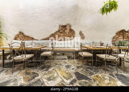 Wooden tables and metal chairs with fabric cushions in a restaurant dining room on a terrace with a blue painted metal pergola and slate floors Stock Photo