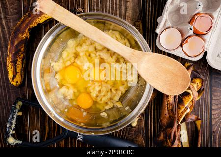Mashed Bananas and Eggs in a Mixing Bowl with a Wooden Spoon: Ingredients for making banana bread with a potato masher and banana peels Stock Photo