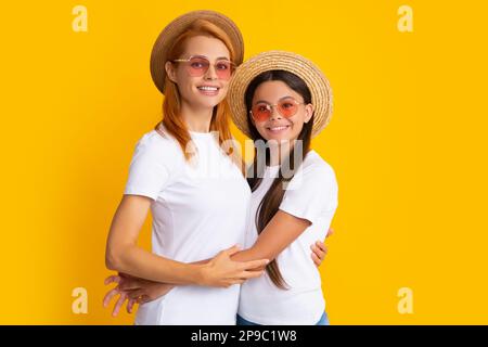 Young mother and teenage girl daughter lovely hugging and cuddling, wearing t-shirt straw hat and sunglasses, isolated on bright yellow background Stock Photo