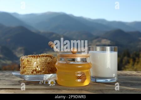 Fresh aromatic honey, combs and glass of milk on wooden table against mountain landscape Stock Photo