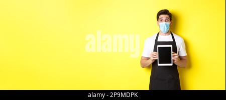 Concept of covid-19, small business and pandemic. Surprised waiter in black apron and medical mask showing tablet screen, standing over yellow Stock Photo