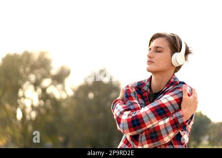 Young urban woman in headphones breathing fresh air and self hugging outdoors. Stock Photo