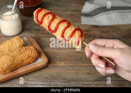 Woman holding delicious corn dog with ketchup at wooden table, closeup Stock Photo