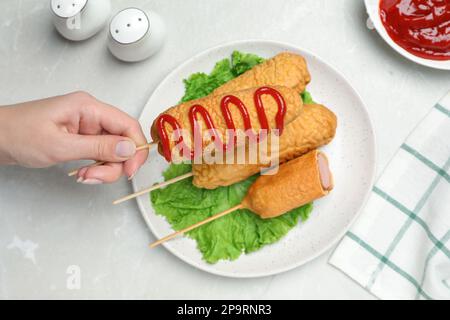 Woman holding delicious corn dog with ketchup at light table, top view Stock Photo