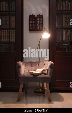 Comfortable armchair with book and lamp between wooden bookcases in library Stock Photo