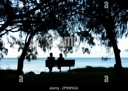 A rear view of a couple sitting on a wooden bench on the shore of Torquay beach, Hervey Bay, Queensland, Australia Stock Photo