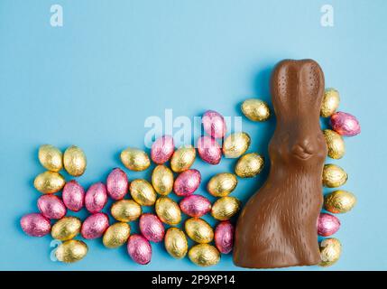 Big milk chocolate bunny and pile of Easter candy eggs foil wrapped in pink and golden on blue background. Happy Easter concept. Preparation for holid Stock Photo