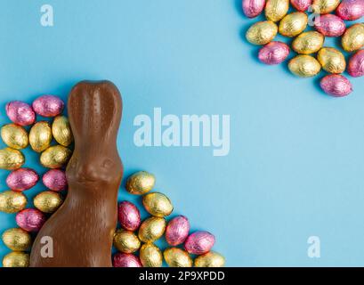 Big milk chocolate Easter bunny and frame of pile of candy eggs wrapped in pink and golden foil on blue background. Happy Easter concept. Preparation Stock Photo