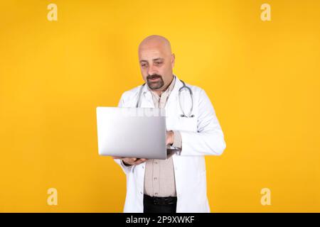 Doctor using laptop. Standing isolated yellow background. Holding his notebook computer, typing. Wearing white uniform, coat with stethoscope. Stock Photo