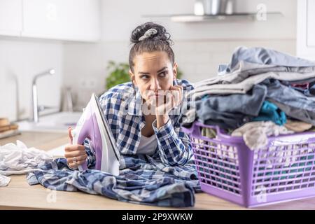 Housewife reluctantly looks at the pile of clothes that need ironing with an iron in her hand. Stock Photo