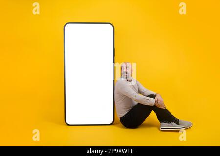 Caucasian millennial guy sitting near big cell phone with empty blank screen. Looking to smartphone. Using mobile devices concept idea. Stock Photo