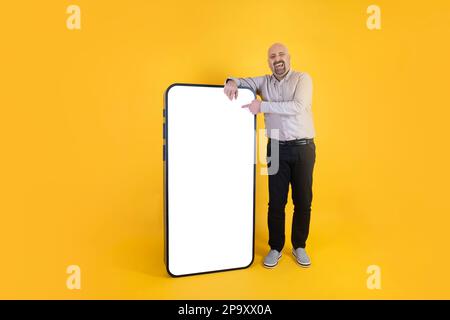 Laughing caucasian man leaning big smartphone. Pointing funny picture or video on social media application. Empty white blank screen for mock up. Stock Photo