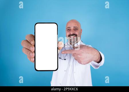 Portrait of excited doctor holding big smartphone pointing white blank screen for mock up. Looking camera. Blue background and copy space. Stock Photo