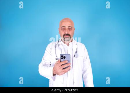Man using modern smartphone, caucasian doctor standing over blue background. Modern bald male hospital professional typing sms message, browsing. Stock Photo
