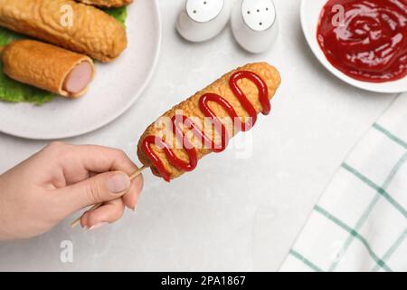 Woman holding delicious corn dog with ketchup at light table, closeup Stock Photo