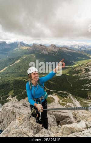 Young attractive female mountain climber on a steep and exposed Via Ferrata in Alta Badia in the South Tyrol in the Italian Dolomites pointing to the Stock Photo