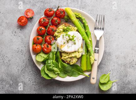 Healthy vegetarian meal plate. Toast, avocado, poached egg, asparagus, baked tomatoes, spinach. Stone background. Vegetarian breakfast plate. Clean Stock Photo