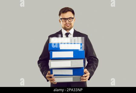 Young displeased Caucasian business man holds lot of folders for documents stands in gray studio Stock Photo