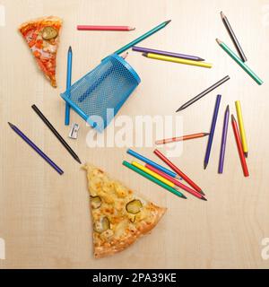 Different colored wooden pencils with metal holder, sharpener and two pieces of pizza scattered haphazardly on wooden table close up Stock Photo