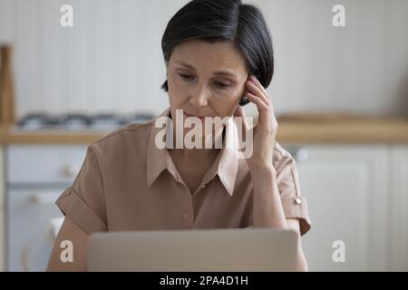 Serious mature freelancer woman using laptop computer in home kitchen Stock Photo