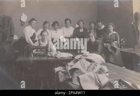 Early 20th century photograph of a group of women workers in a textile workshop. Two women are sitting at treadle sewing machines. The others are standing in the background. A large pile of cloth is in the foreground of the picture and a jacket is hanging up on the left side. Stock Photo