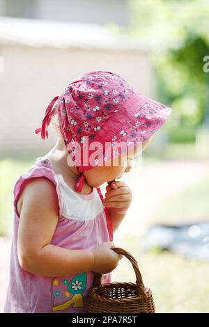 A little girl in a panama eats fresh cherry berries from a basket in garden tree Stock Photo