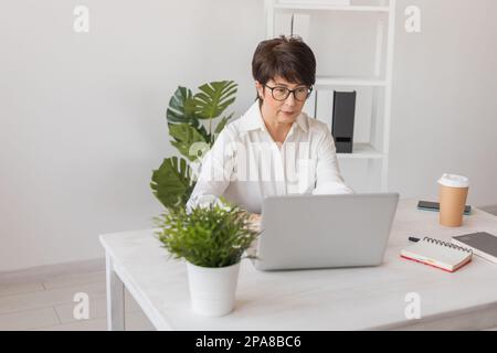 Portrait of beautiful middle aged brunette businesswoman sitting at light modern work place office and typing on laptop Stock Photo