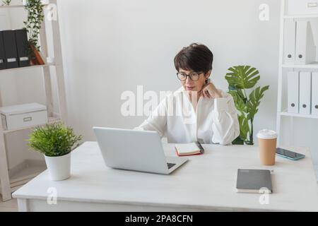 Portrait of beautiful middle aged brunette businesswoman sitting at light modern work place office and typing on laptop Stock Photo