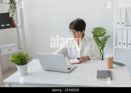 Portrait of beautiful middle aged brunette businesswoman sitting at light modern work place office and typing on laptop Stock Photo