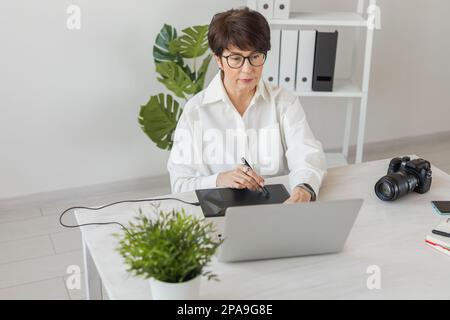 Middle aged woman graphic designer or photographer using digital graphic tablet while working at modern office, professional female retoucher sitting Stock Photo