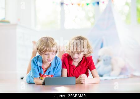 Kids room with teepee tent. Children play computer game on tablet device in white sunny bedroom. Cozy light interior for kid nursery or playroom. Stock Photo