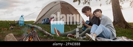 Man playing guitar for woman at fire and tent at nature. Romantic evening. Stock Photo