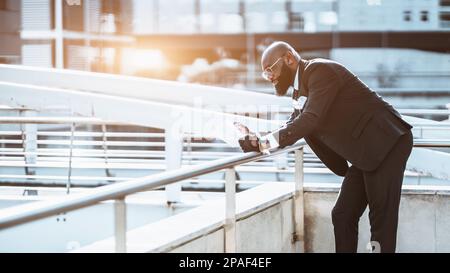 View of a fancy mature bald black afro man in a tailored full black suit, eyeglasses and tie leaning on the railing for better reading the paperwork t Stock Photo