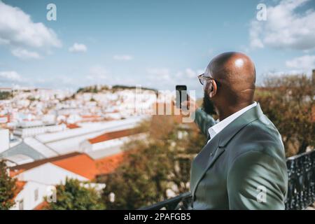 Capture in the selective focus a bald, bearded afro tourist man in a pastel green suit jacket takes pictures of the city from a viewpoint on his smart Stock Photo