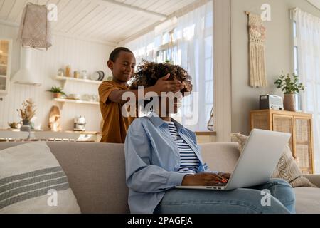 Playful kid son covering eyes of smiling surprised mixed race curly mother have fun together at home Stock Photo