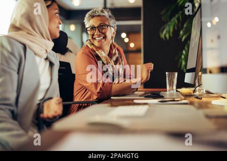 Two happy businesswomen having a discussion while working at their desks in an office. Diverse businesswomen working as a team in a creative co-workin Stock Photo