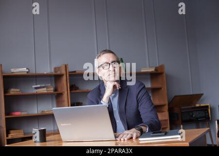 Portrait of grey haired pensive businessman in glasses and blue suit sitting and looking on laptop Stock Photo