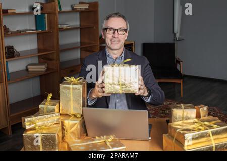 Mature happy smilimg  businessman in glasses and blue suit with laptop and a lot of gifts around. He is holding gold box with gold ribbons Stock Photo
