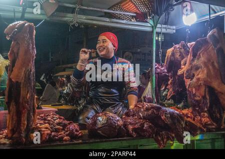 A happy Cambodian woman eats a skewer and sells raw meat at the main wholesale meat market, Phsar Dumkor, at night. Phnom Penh, Cambodia. © Kraig Lieb Stock Photo