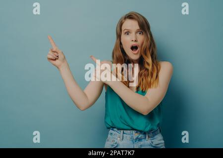 Surprised young female in casual wear pointing to right side with index fingers shocked by what she saw and looking at camera with mouth wide open iso Stock Photo