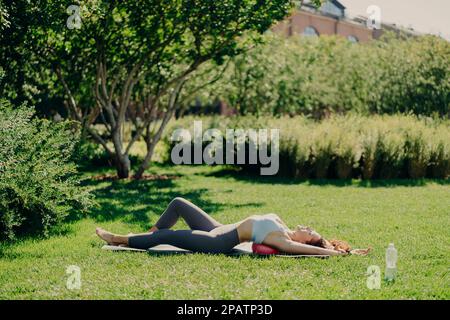 Relaxed sporty woman lying on fitness ball at karemat does aerobics exercises wears cropped top and leggings breathes fresh air deeply has perfect fig Stock Photo