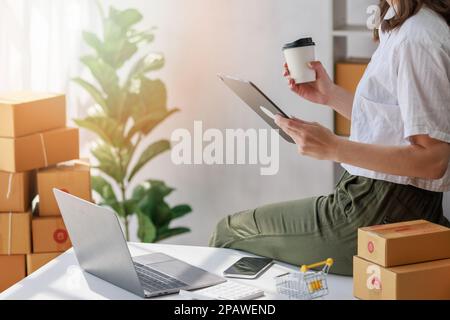Concept of parcel delivery and selling online,Retailer Using computer tablet to check sales for the day on an online store while holding coffee cup wi Stock Photo