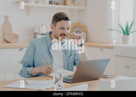 Smiling male employee using laptop at home sitting at kitchen table, looking at screen and making video call or watching webinar, writing notes in not Stock Photo