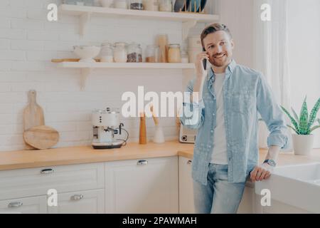 Handsome smiling young bearded man wearing casual clothes talking on mobile phone in kitchen while working online remotely from home, having successfu Stock Photo