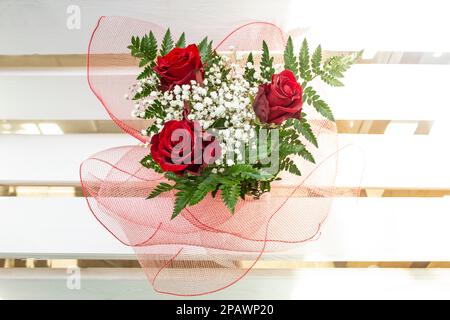 Three roses bouquet, with red wire mesh folds, on white horizontal wood boards, top view close up. Stock Photo